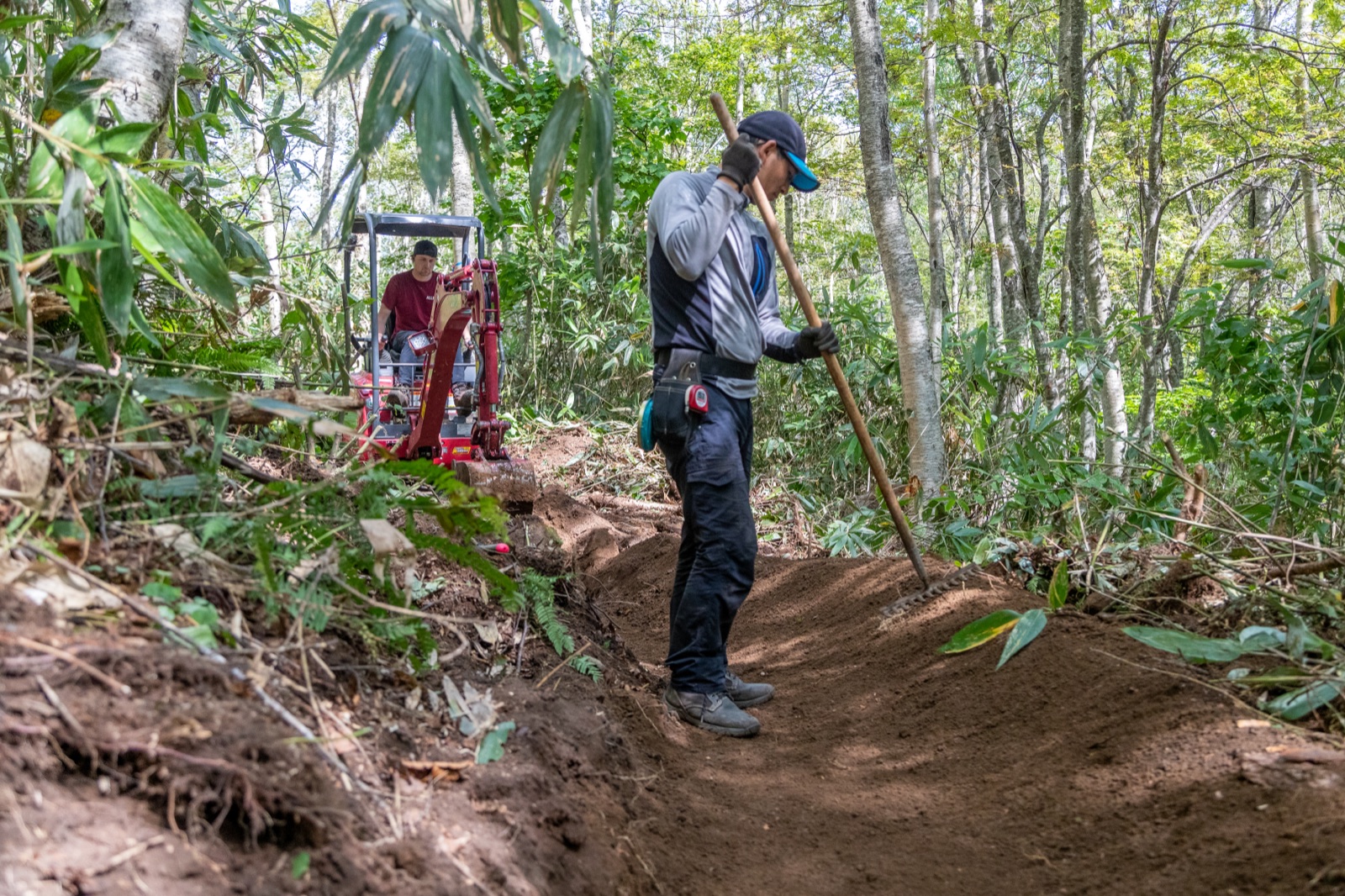 Trail crew hand-finishing a berm with a mattock while a mini-excavator works behind in the forest