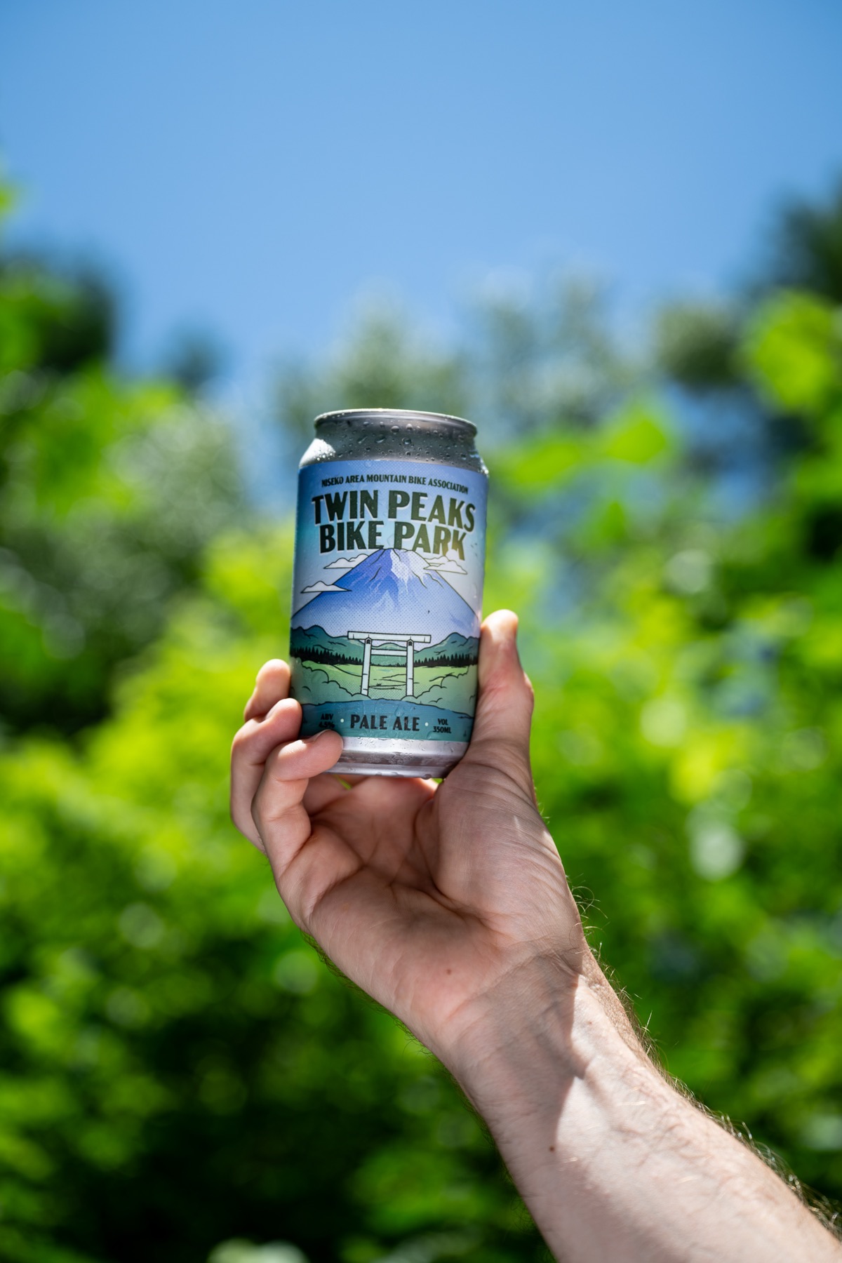 A hand holding up a can of Twin Peaks Bike Park Pale Ale against summer foliage, showing Joe Scott's label artwork of Mount Yotei above the park's torii gate