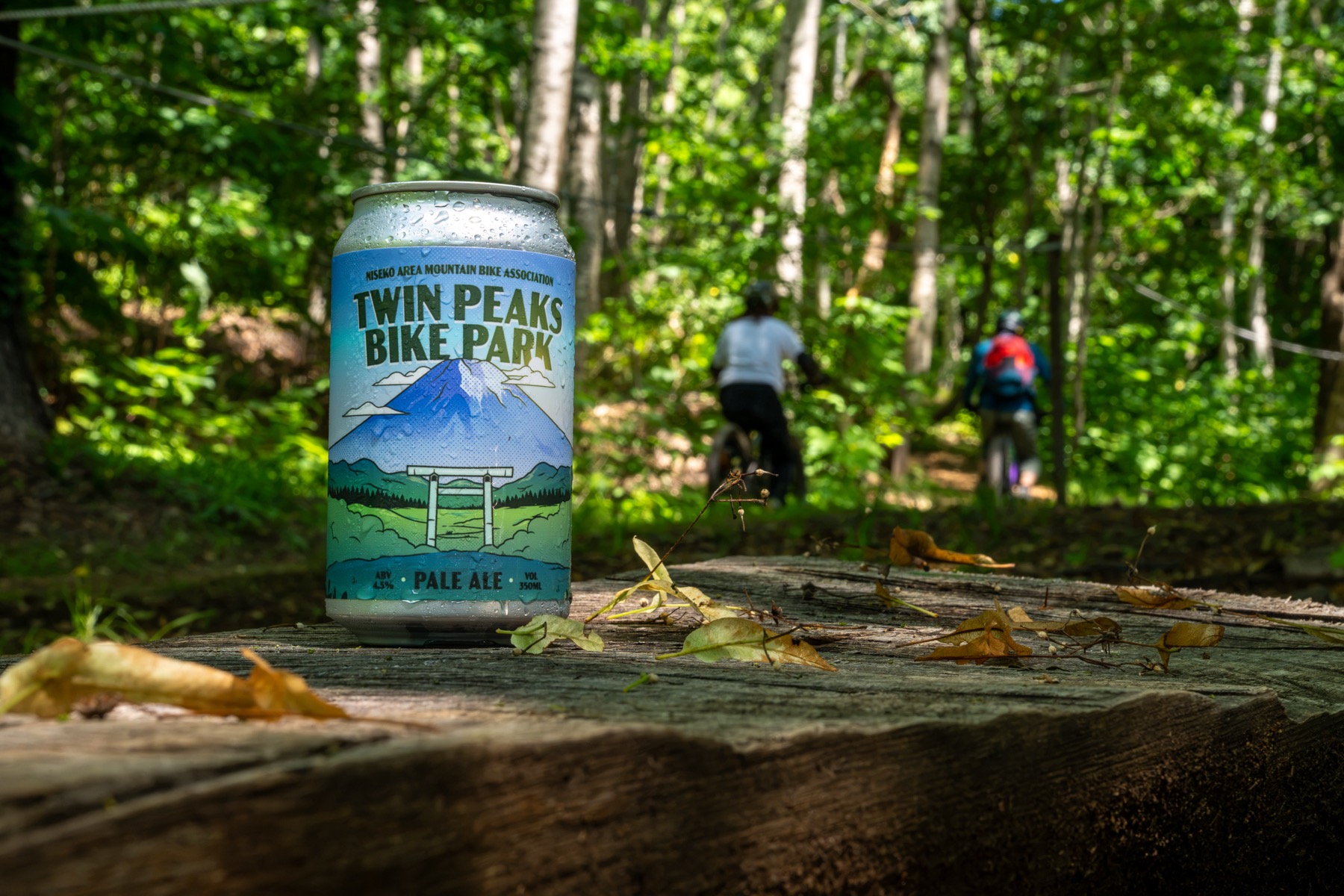 A can of Twin Peaks Bike Park Pale Ale resting on a log as two riders pass on the trail behind