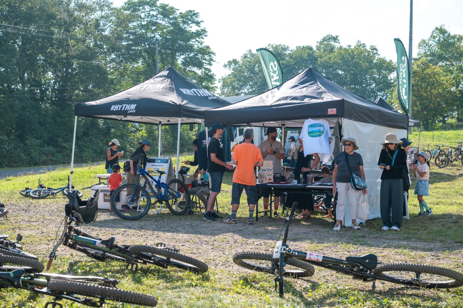 Two bike shop tents pitched on grass, with riders and families looking over gear laid out on tables