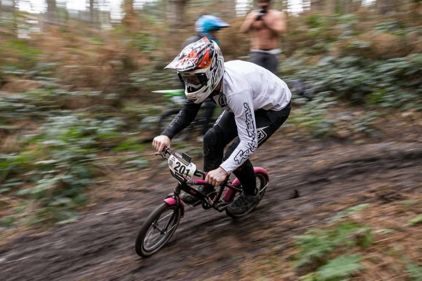 An adult rider in a full-face helmet racing down a dirt track on a tiny kids' BMX