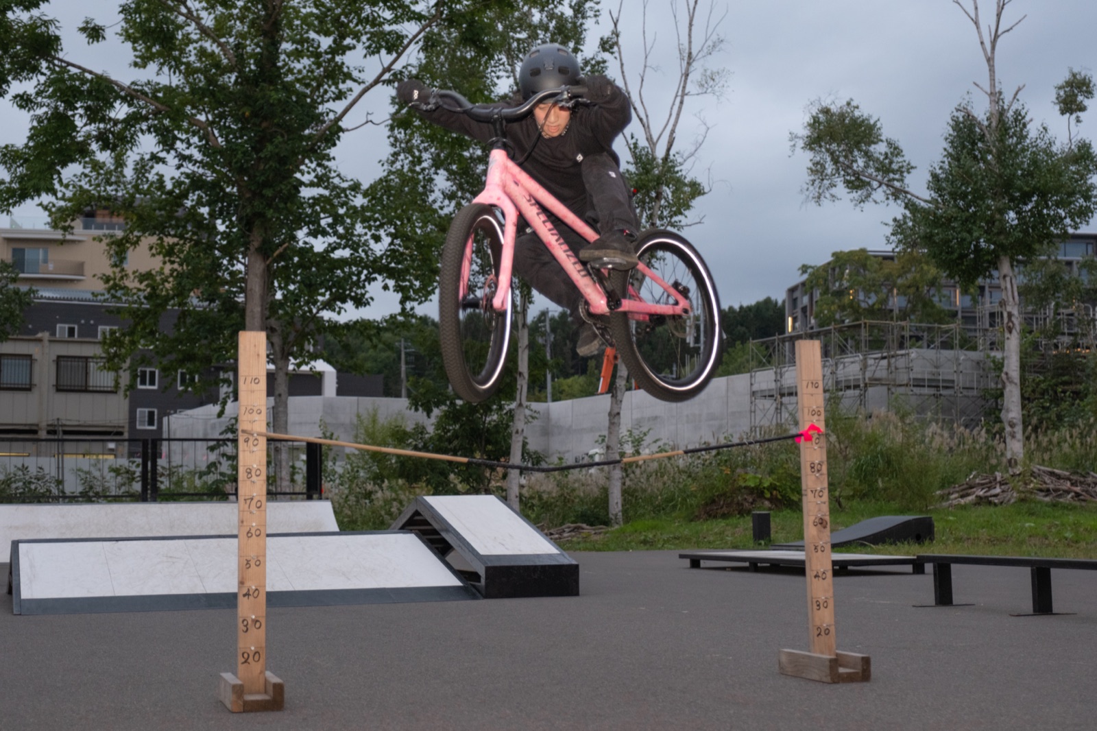 A rider in mid-air clearing an elastic bar strung between two measured wooden posts at a bike park