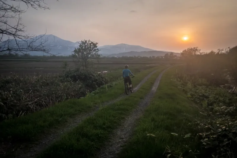 夕暮れの​グラベルロードを​走る​ライダー、​遠くに​ニセコ連峰の​山並み