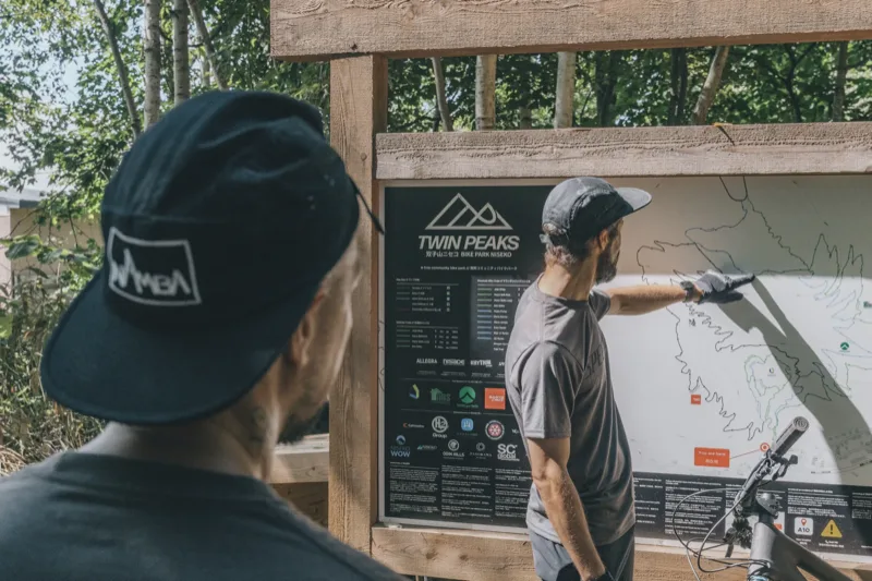 Two riders looking at the Twin Peaks Bike Park trail map board, sponsor logos visible along the bottom