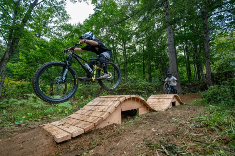 ツインピークススキルセンターで​ウッドフィーチャーを​越える​ライダー、​白樺の​森が​背景