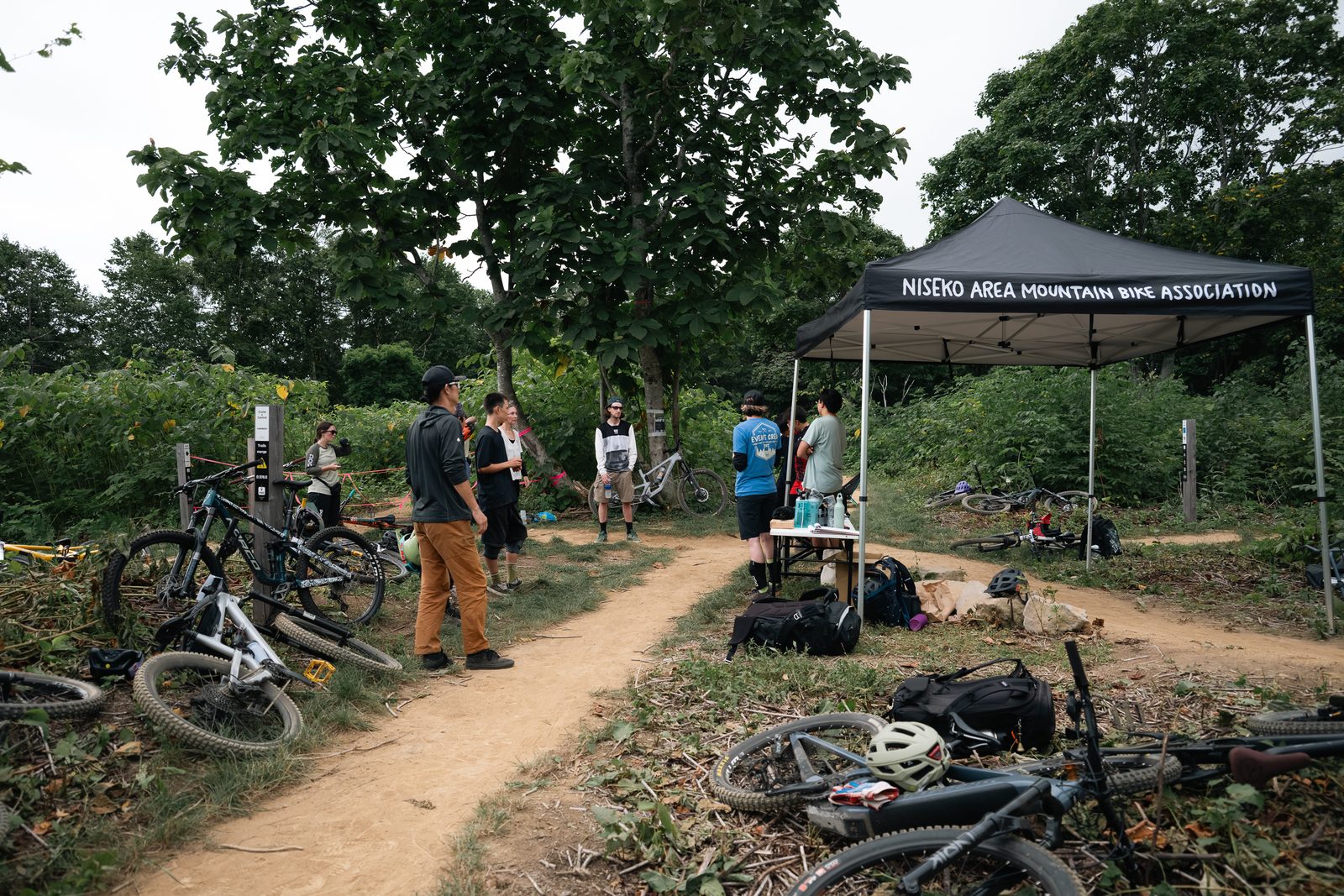 夏の白樺林、トレイル上部に張られたNAMBAのイベントテント。地面にバイクが横たわり、数人が集まっている