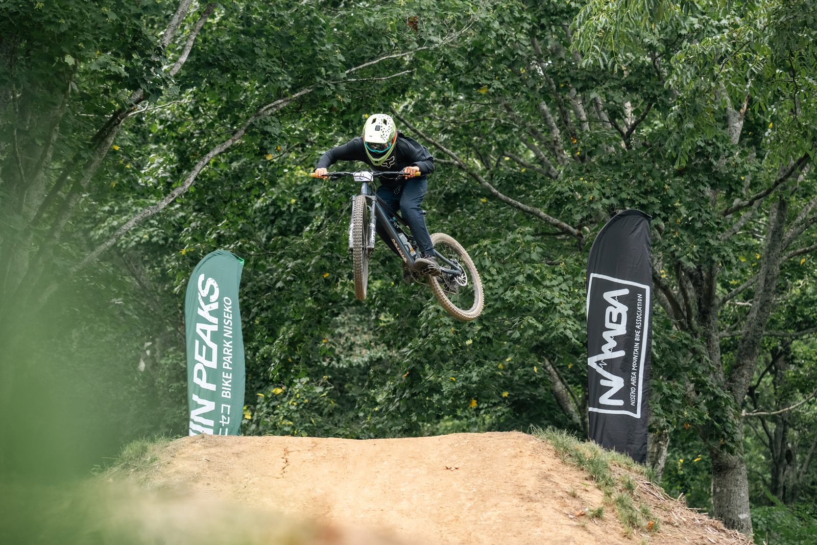 ライムグリーンのヘルメットとネイビーのトップスのライダーが、ビッグジャンプのリップを越えながらバイクを横に振る