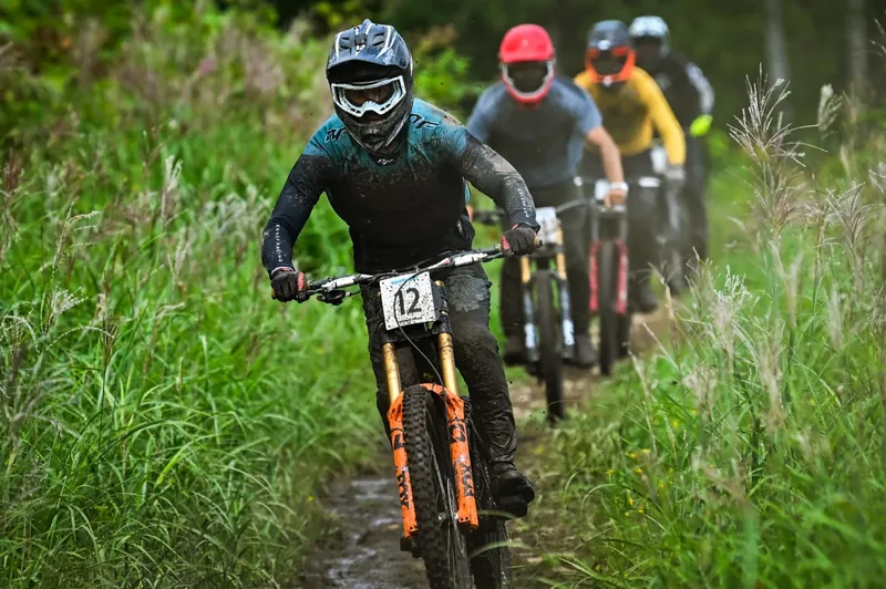 Four downhill racers strung out along a muddy, grass-lined section of the Annupuri course