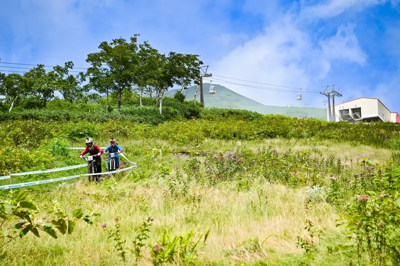 Two riders push their bikes up through a wildflower meadow with Mount Annupuri and the gondola line behind