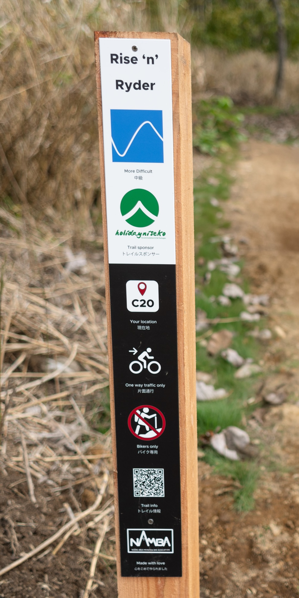Close-up of a NAMBA trail sign for Rise 'n' Ryder showing trail name, difficulty, sponsor, location code, direction, trail users, QR code, and NAMBA logo